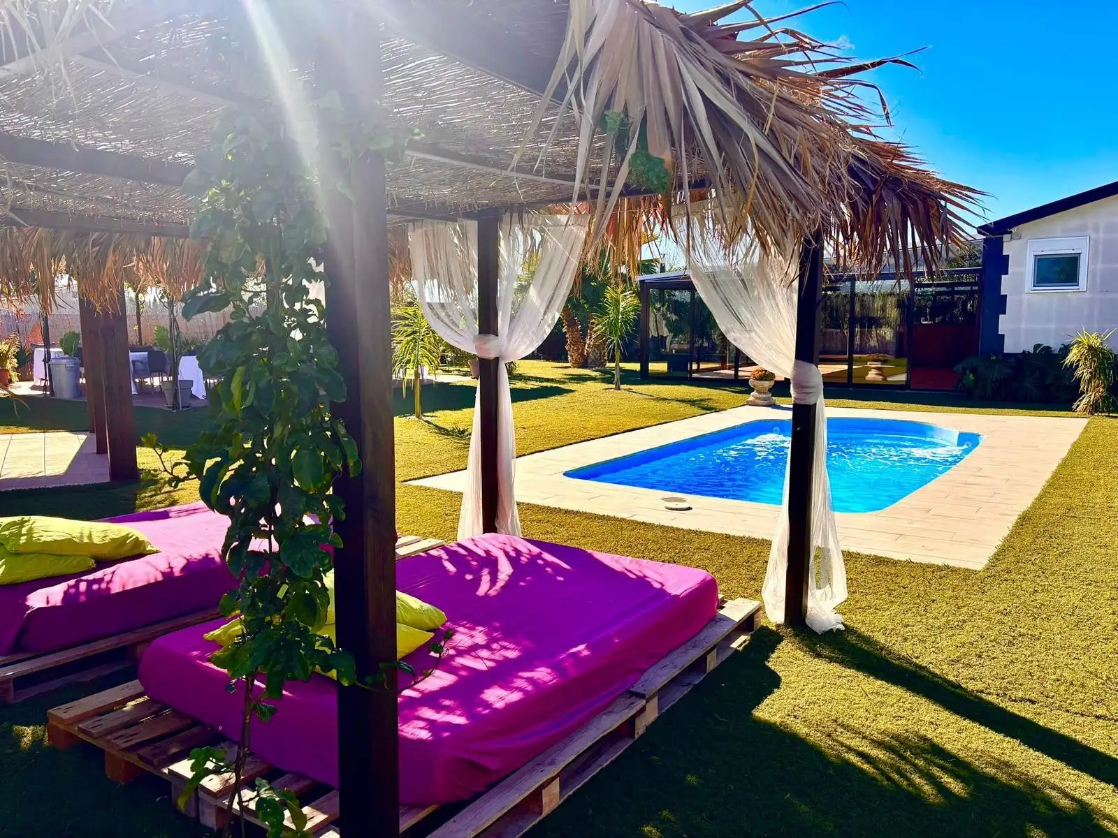 La luz del sol atravesando una cabaña de palma con cortinas, camas fucsia y la piscina detrás