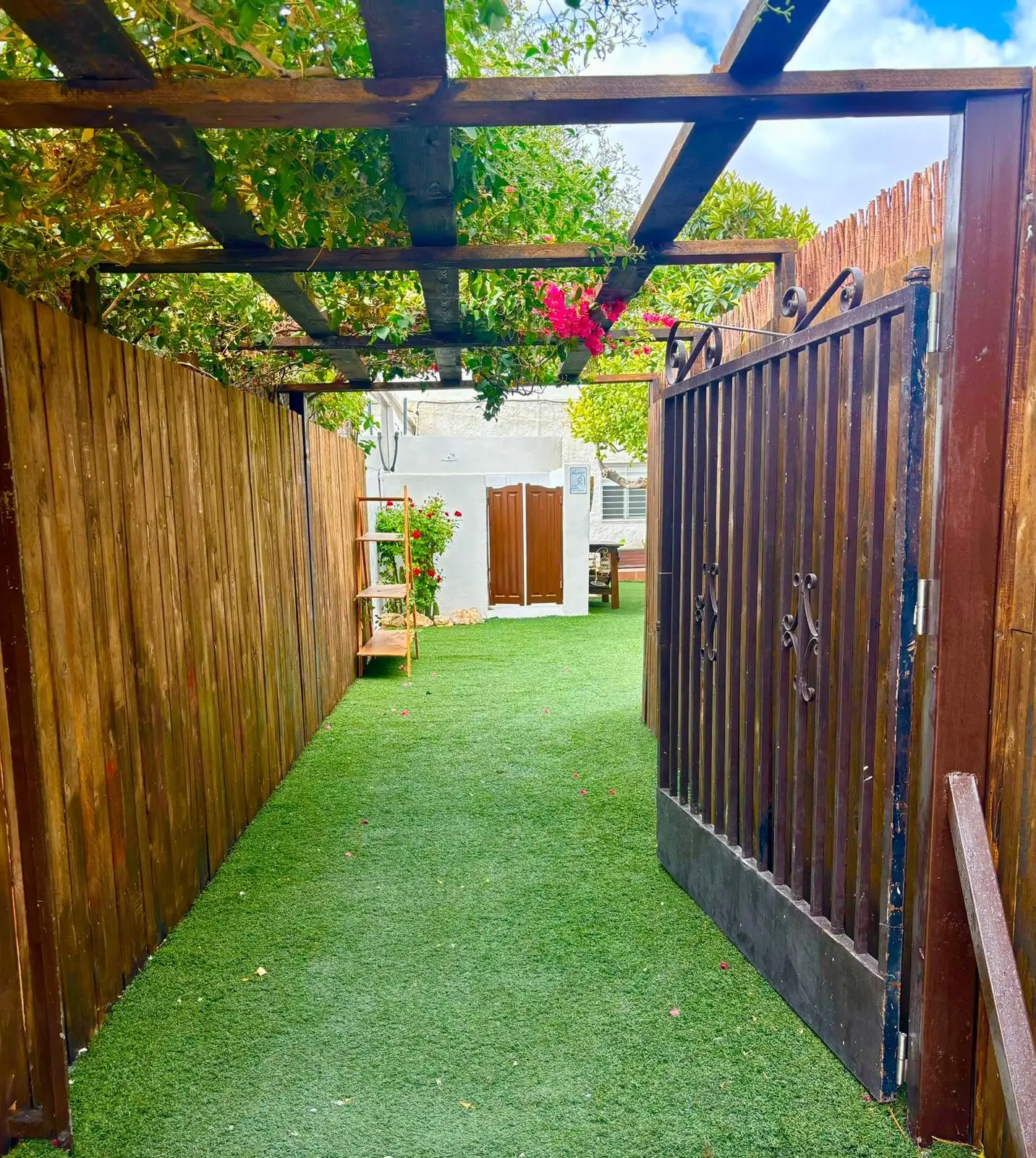 Un pasillo de césped entre una valla de madera y un muro blanco, con un portón de hierro a la derecha y una pérgola con parra por encima
