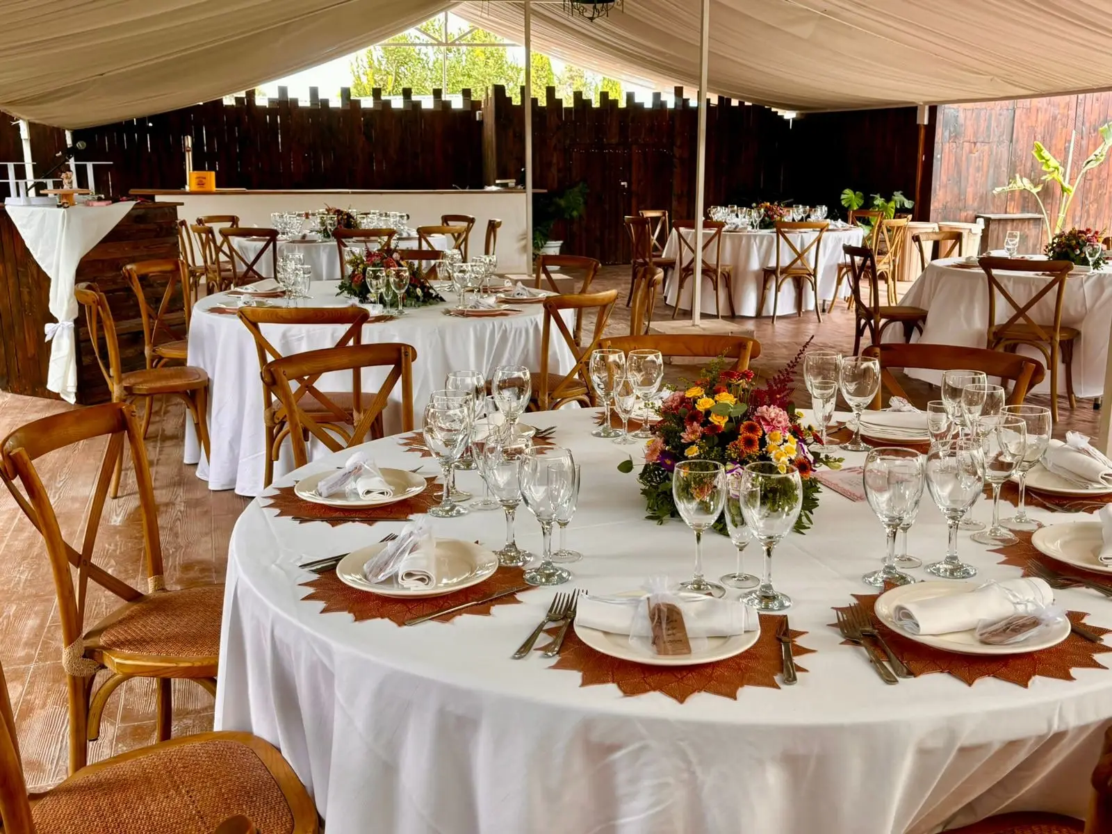 Bajo la carpa, mesas redondas con mantel blanco, cubiertos, copas y un centro de flores, con sillas de madera