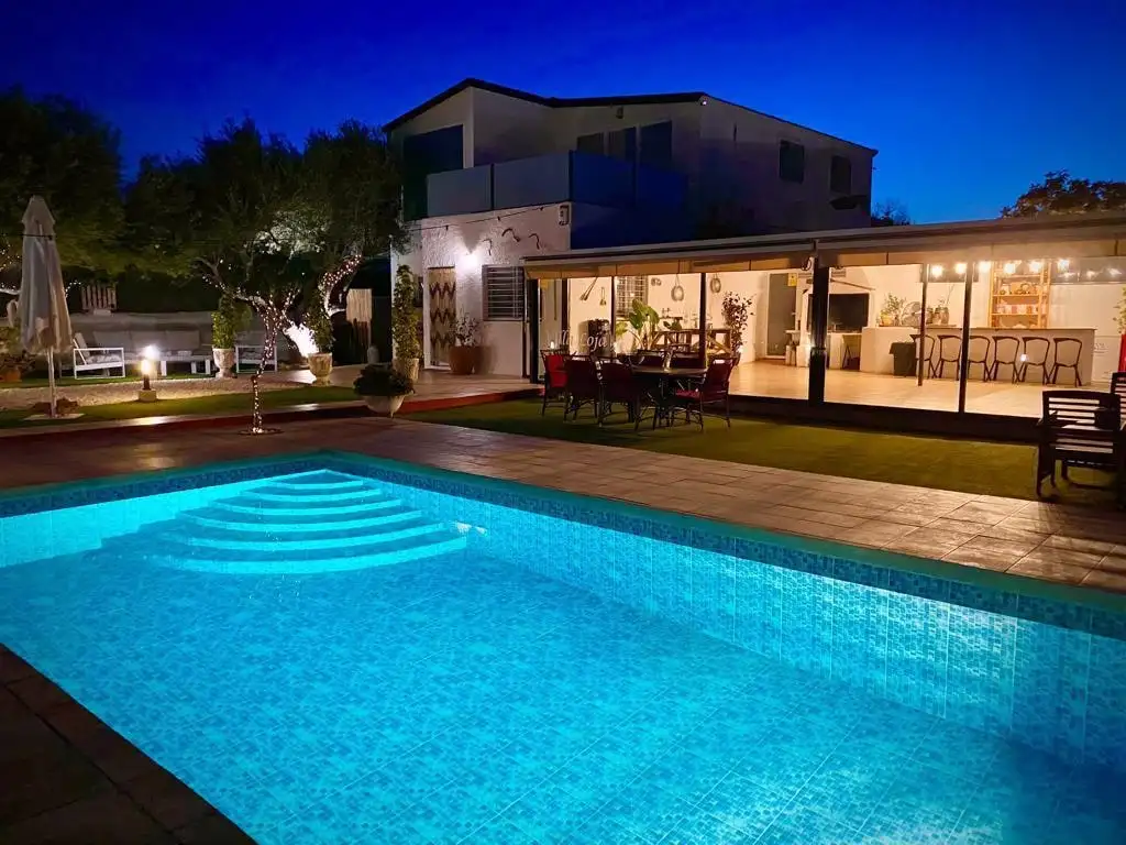 La casa de dos plantas de noche, con la piscina iluminada en azul y el salón encendido