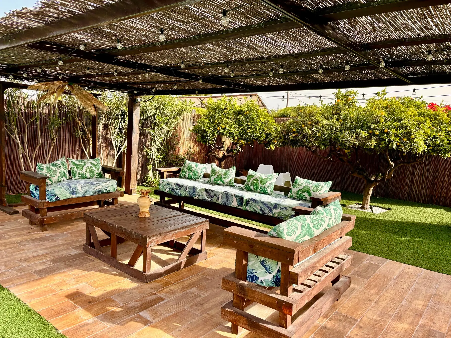 Bajo una pérgola de cañizo, sofás de palés con cojines de hojas tropicales alrededor de una mesa baja