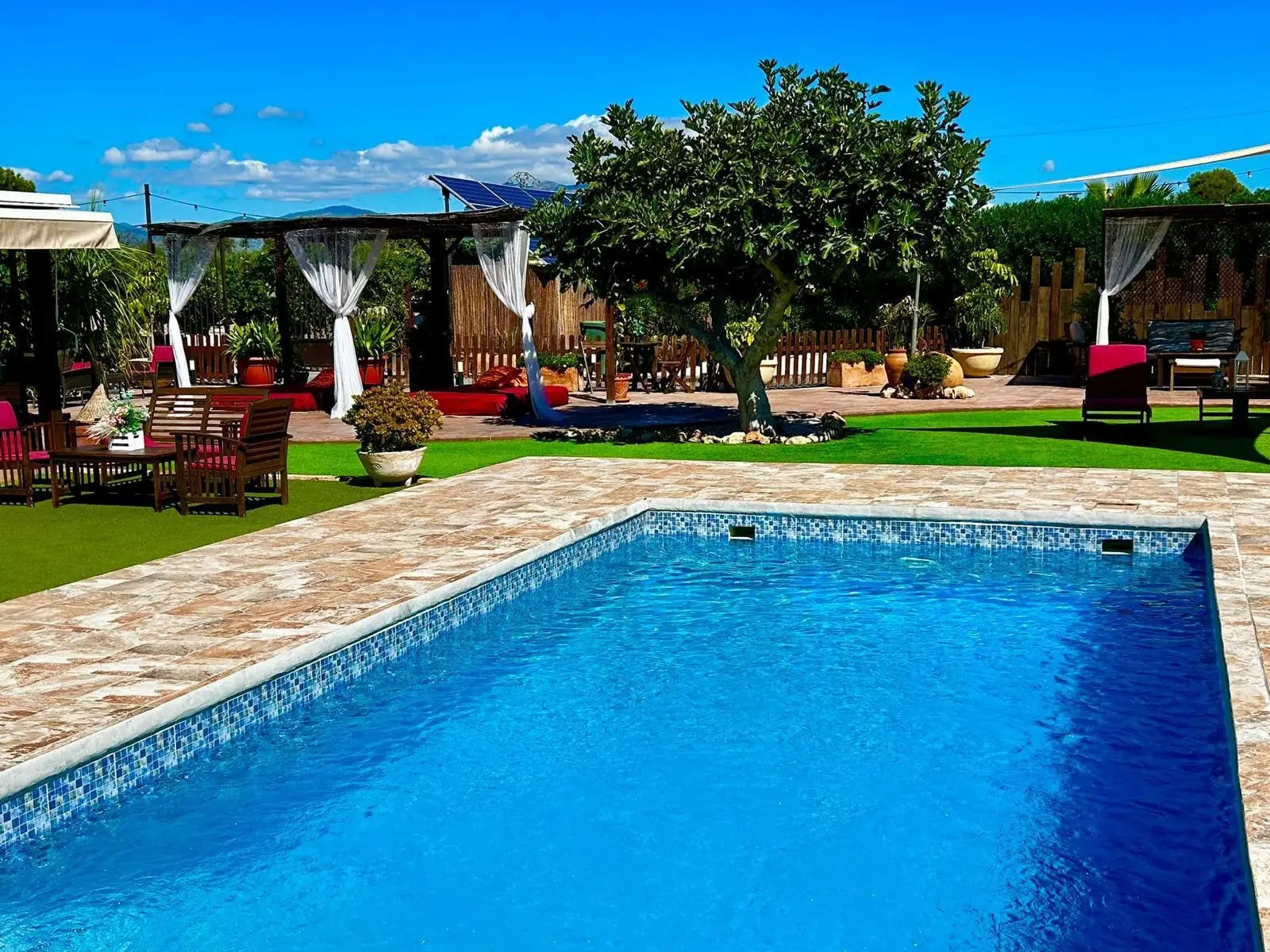 La piscina, una higuera y las pérgolas con cortinas blancas al fondo, con las montañas en el horizonte