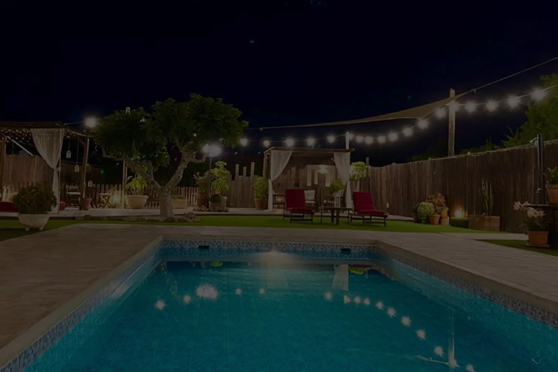 Toda la piscina de noche, con guirnaldas de luces sobre el jardín y las cabañas con cortinas al fondo
